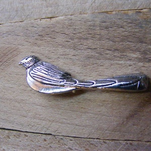 May include: A silver bird brooch with detailed wings and a long tail. The bird is facing left and has a slightly curved beak.