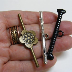 May include: A collection of metal toggle clasps in various designs. Includes a bronze-colored flower-shaped clasp, a silver-colored bar clasp, and a black, textured bar clasp with a ring. The clasps are held in a hand against a white background.