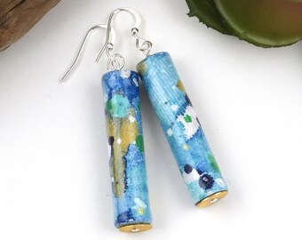 May include: A pair of dangle earrings with blue and gold painted wooden cylinder beads. The beads have a speckled pattern and are attached to silver hooks.