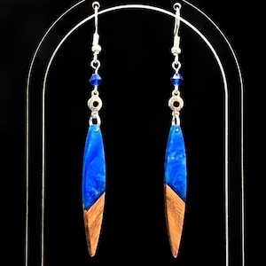 May include: A pair of dangle earrings with a blue resin and wood design. The earrings have silver-colored hooks and are accented with blue beads. The bottom half of each earring is wood, and the top half is a vibrant blue resin.