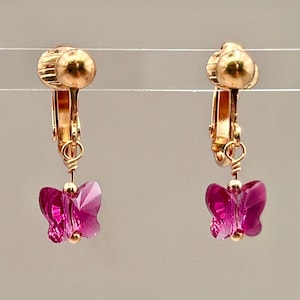 May include: A pair of gold-toned clip-on earrings, each featuring a faceted, magenta butterfly charm. The butterfly charms are suspended from a gold-colored clip. The earrings are displayed against a neutral background.