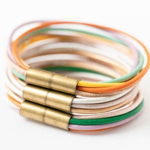 May include: A multi-colored leather bracelet with a brass clasp. The bracelet is made of several thin strips of leather in shades of orange, yellow, green, purple, and white.