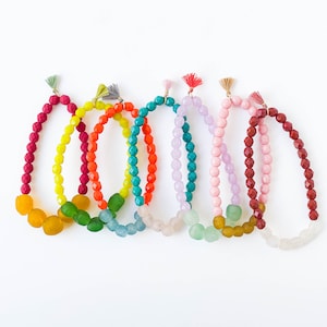 May include: Seven colourful beaded bracelets with tassel accents. The bracelets are made of glass beads in shades of red, orange, yellow, green, blue, purple, and pink.