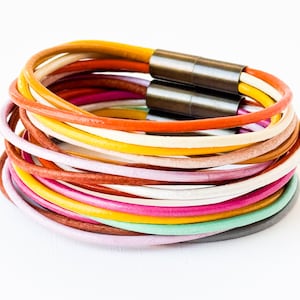 Könnte beinhalten: Mehrfarbiges Lederarmband mit einem silbernen Metallverschluss. Das Armband besteht aus vielen dünnen Ledersträngen in verschiedenen Farben, darunter Gelb, Orange, Rot, Rosa, Lila, Blau, Grün und Braun.