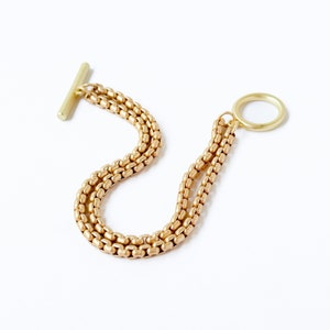 May include: A gold-toned chain bracelet with a T-bar clasp and a circular ring at the other end. The bracelet is made of multiple strands of small, linked beads.