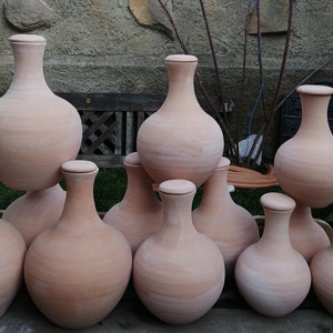 Peut inclure: Dix cruches en terre cuite avec un corps rond et un col étroit. Les cruches sont disposées en quinconce sur une surface en bois.