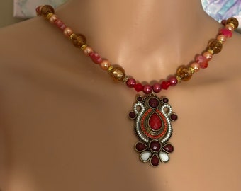 Könnte beinhalten: Eine Perlenkette mit einem großen, verzierten Anhänger. Die Kette besteht aus rosa, orangefarbenen und goldenen Perlen. Der Anhänger ist aus Metall und hat ein rotes, orangefarbenes und weißes Design.