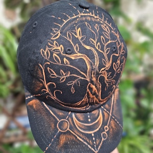 May include: A black baseball cap featuring a hand-painted orange design of a sprawling tree with intricate branches and leaves on the crown, and a geometric pattern with a crescent moon and circular shapes on the curved brim of the hat.