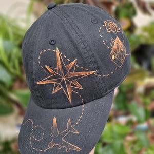 May include: Black baseball cap with a compass rose, airplane, and camera design in a burnt orange color. The cap has a curved brim and a button on top. The design elements are connected by dotted lines.