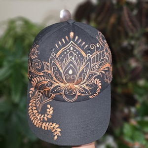 Op de afbeelding: Een zwarte baseballcap met een koperkleurig lotusbloemontwerp op de voorkant. De cap heeft een gebogen klep en een decoratief bloemmotief aan de zijkant. De cap wordt door een hand omhoog gehouden.