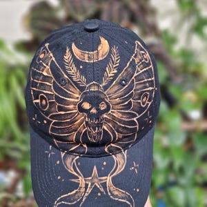 May include: A black baseball cap featuring a bleached design of a moth with a skull-shaped body. Above the moth is a crescent moon and two sprigs of leaves. Below the moth, on the brim, is a star surrounded by small dots and curved lines.
