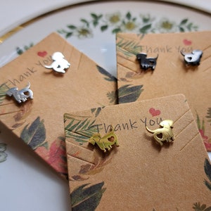 Könnte beinhalten: Dackel-Ohrstecker in Silber, Schwarz und Gold. Jedes Paar wird auf einer braunen Karte mit floralem Design, den Worten "Thank You" und einem roten Herz präsentiert. Die Ohrringe liegen auf einem weißen Teller.