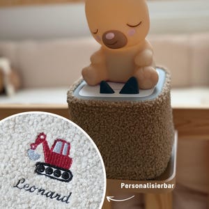 Könnte beinhalten: Ein beigefarbenes Bären-Nachtlicht mit geschlossenen Augen sitzt auf einem braunen, plüschigen Sockel. Der Sockel hat eine weiße Oberseite mit zwei schwarzen Dreiecken. Eine Nahaufnahme zeigt einen gestickten roten Bagger und den Namen "Leonard" auf weißem Stoff.