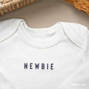 Könnte beinhalten: Cremefarbener Baby-Body mit dem Wort "NEWBIE" in marineblauen Blockbuchstaben auf der Vorderseite aufgedruckt. Der Body hat einen runden Halsausschnitt und lange Ärmel. Der Stoff wirkt weich und bequem.