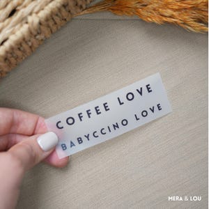 Könnte beinhalten: Ein rechteckiges, durchscheinendes Etikett mit den Wörtern "COFFEE LOVE" und "BABYCCINO LOVE" in Dunkelblau gedruckt. Das Etikett wird von einer Hand mit lackierten Nägeln vor einem neutralen Hintergrund gehalten.