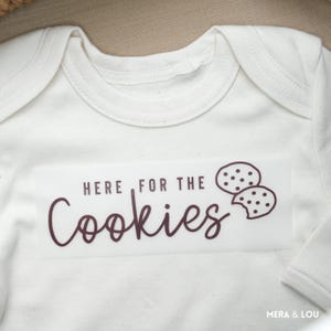 Könnte beinhalten: Cremefarbener Baby-Body mit dem Schriftzug "HERE FOR THE Cookies" in dunkelbrauner Schrift. Das Design beinhaltet zwei Keks-Illustrationen, eine davon mit einem Biss. Der Body ist aus weichem Stoff.