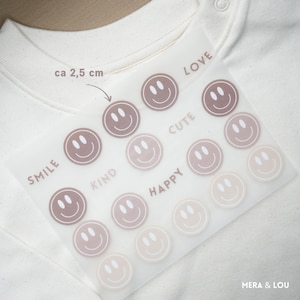 Könnte beinhalten: Ein Bogen mit Smiley-Aufklebern in verschiedenen Braun- und Rosatönen, mit Wörtern wie "Smile", "Kind" und "Happy". Die Aufkleber befinden sich auf einem transparenten Bogen, mit einer Größe von ca. 2,5 cm. Der Markenname "MERA & LOU" ist sichtbar.