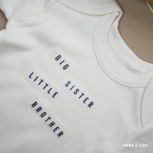 Könnte beinhalten: Cremefarbener Baby-Body mit den Wörtern "BIG", "LITTLE", "SISTER" und "BROTHER" in Schwarz aufgedruckt. Der Body besteht aus weichem Stoff und hat einen runden Halsausschnitt.