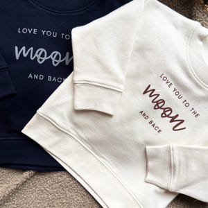 Könnte beinhalten: Zwei Sweatshirts, eines in Marineblau und eines in Creme, werden gezeigt. Das marineblaue Sweatshirt hat den Text "LOVE YOU TO the moon AND BACK" in Silber. Das cremefarbene Sweatshirt hat den gleichen Text in Braun. Beide haben lange Ärmel.