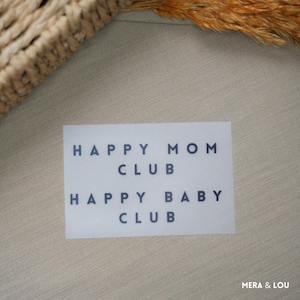 Könnte beinhalten: Eine weiße rechteckige Karte mit dem Text "HAPPY MOM CLUB" und "HAPPY BABY CLUB" in dunkelblauer, serifenloser Schrift. Die Karte liegt auf einer hellbeigen Oberfläche, mit einem geflochtenen Korb und getrockneten Pflanzenstängeln im Hintergrund.