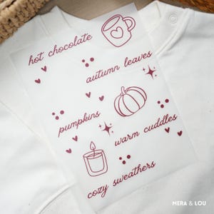 Può includere: Una felpa bianca con una sovrapposizione trasparente con illustrazioni e testo a tema autunnale. La sovrapposizione include una tazza con un cuore, una zucca, una candela e le parole "cioccolata calda", "foglie d'autunno", "zucche", "caldi abbracci" e "maglioni accoglienti".