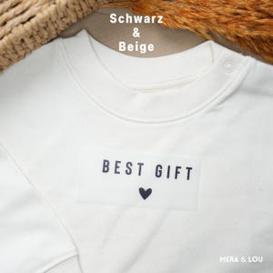 Könnte beinhalten: Cremefarbenes Sweatshirt mit dem schwarzen Schriftzug "BEST GIFT" und einem kleinen schwarzen Herz. Das Sweatshirt hat einen runden Ausschnitt und einen Druckknopfverschluss. Der Markenname "MERA & LOU" ist unten rechts sichtbar.