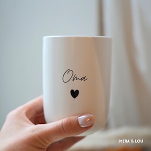 Könnte beinhalten: Eine weiße Keramik-Tasse mit dem Wort "Oma" in schwarzer Schrift und einem kleinen schwarzen Herz. Die Tasse hat eine glatte, matte Oberfläche und ein schlichtes Design. Die Tasse wird von einer Person gehalten.