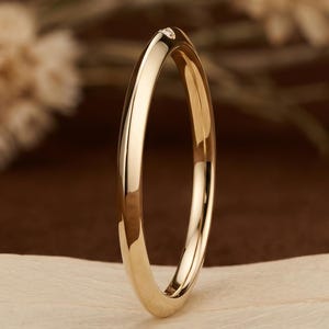 Puede incluir: Una alianza de boda dorada con un pequeño diamante en la parte superior. El anillo se muestra sobre una hoja de color claro sobre un fondo marrón. El logotipo de Luxcraft Jewels es visible en la esquina inferior derecha.