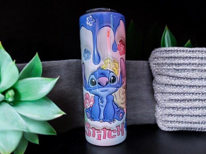 May include: A blue and white stainless steel tumbler with a colorful design featuring Stitch from Lilo & Stitch. The design includes flowers, leaves, and the word "Stitch" in pink letters.