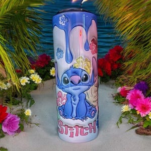 May include: A blue and white stainless steel tumbler with a cartoon image of Stitch from Lilo & Stitch. The tumbler has a dripping paint design and says "Stitch" at the bottom.