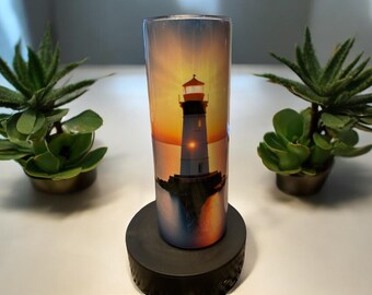 May include: A stainless steel tumbler with a colorful image of a lighthouse at sunset. The tumbler is on a black coaster.