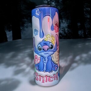 May include: A blue and white stainless steel tumbler with a cartoon image of Stitch from Lilo & Stitch. The tumbler has a pink, blue, and yellow floral design and the word "Stitch" is written in pink letters at the bottom.