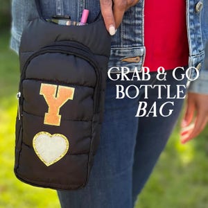 May include: Black quilted bottle bag with a gold glitter heart and a gold letter "Y". The bag has a small zippered pocket and a pocket for a bottle. The text "GRAB & GO BOTTLE BAG" is printed on the right side.
