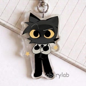May include: A clear acrylic keychain featuring a cartoon character with a black cat head, large yellow eyes, and a white shirt with a black vest and trousers. The character has arms crossed and is surrounded by two small yellow stars. The text Kaborylab is visible.