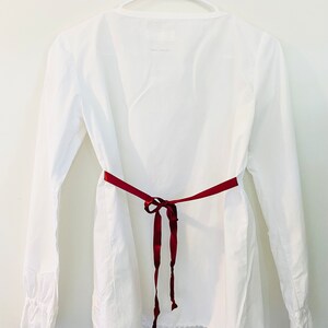 May include: A white long-sleeved blouse with a V-neck and a burgundy ribbon tied at the waist. The blouse has a scalloped hem and is hanging on a white hanger against a white wall.
