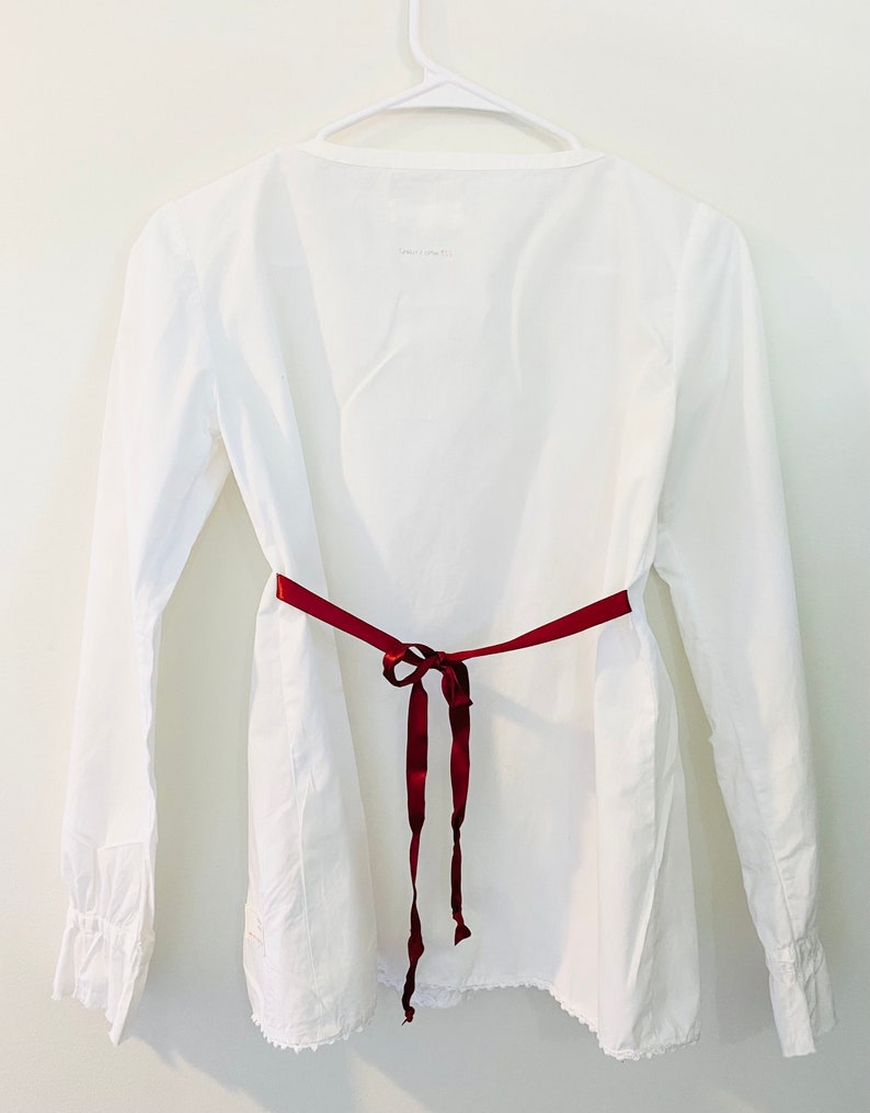 May include: A white long-sleeved blouse with a V-neck and a burgundy ribbon tied at the waist. The blouse has a scalloped hem and is hanging on a white hanger against a white wall.