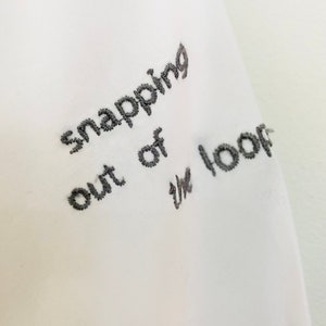 May include: Close-up of white fabric with embroidered text in dark gray. The text reads "snapping out of the loop". The embroidery is detailed and the fabric appears to be a soft material.
