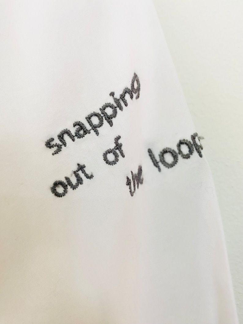 May include: Close-up of white fabric with embroidered text in dark gray. The text reads "snapping out of the loop". The embroidery is detailed and the fabric appears to be a soft material.