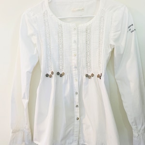 May include: A white button-down shirt with long sleeves, a round neckline, and decorative lace panels down the front. The shirt features small, decorative buttons and embroidered accents. The sleeves have ruffled cuffs. The text "shopping of the" is on the right sleeve.
