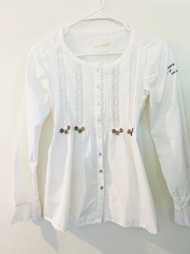 May include: A white button-down shirt with long sleeves, a round neckline, and decorative lace panels down the front. The shirt features small, decorative buttons and embroidered accents. The sleeves have ruffled cuffs. The text "shopping of the" is on the right sleeve.
