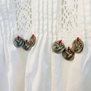 May include: Close-up of a white garment with vertical pleats and a decorative lace panel. Silver-colored dove-shaped charms are attached with red thread. The garment has a vintage or handcrafted aesthetic.