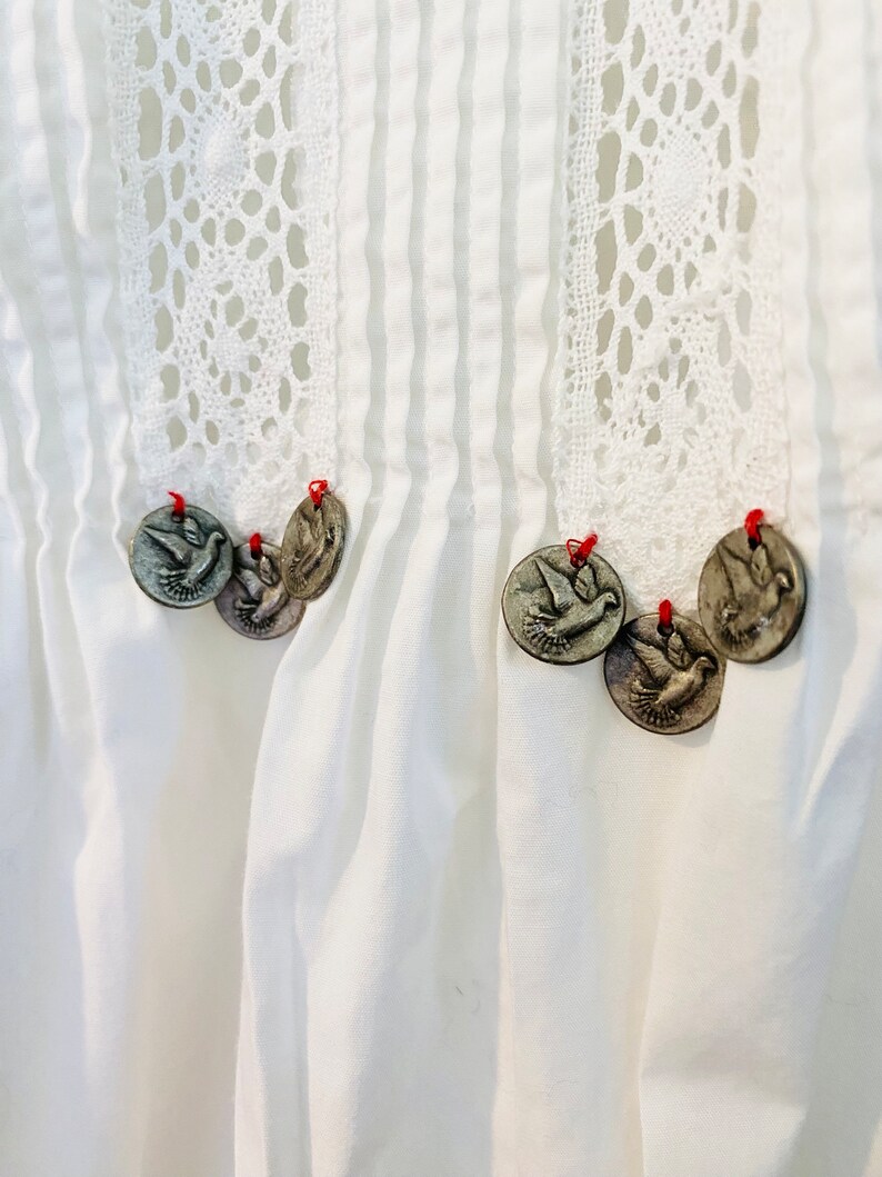 May include: Close-up of a white garment with vertical pleats and a decorative lace panel. Silver-colored dove-shaped charms are attached with red thread. The garment has a vintage or handcrafted aesthetic.