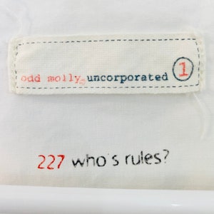 May include: White fabric with embroidered text. The top section reads "odd molly, incorporated" with a red "1" inside a blue circle. The bottom section has "227 who's rules?" in red and black.
