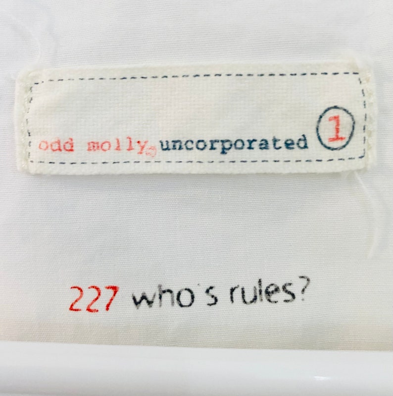 May include: White fabric with embroidered text. The top section reads "odd molly, incorporated" with a red "1" inside a blue circle. The bottom section has "227 who's rules?" in red and black.
