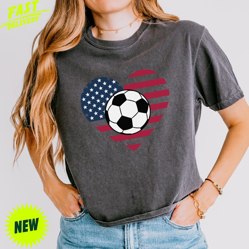 Puede incluir: Una camiseta de manga corta en color gris carb&oacute;n con un gr&aacute;fico en forma de coraz&oacute;n con los colores de la bandera estadounidense y un bal&oacute;n de f&uacute;tbol blanco y negro centrado. La prenda tiene cuello redondo y un corte holgado con aspecto desgastado.