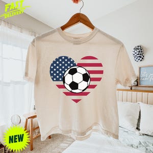 Puede incluir: Una camiseta de manga corta de color crema con un gr&aacute;fico en forma de coraz&oacute;n con el patr&oacute;n de la bandera estadounidense, compuesto por estrellas blancas sobre fondo azul y rayas rojas y blancas, con un bal&oacute;n de f&uacute;tbol blanco y negro en el centro.