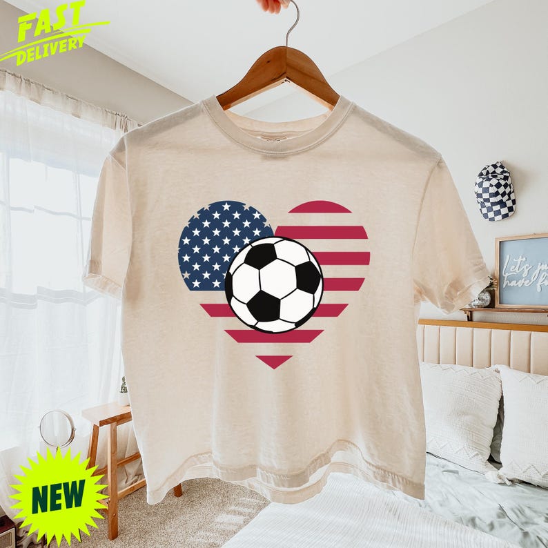 Puede incluir: Una camiseta de manga corta de color crema con un gr&aacute;fico en forma de coraz&oacute;n con el patr&oacute;n de la bandera estadounidense, compuesto por estrellas blancas sobre fondo azul y rayas rojas y blancas, con un bal&oacute;n de f&uacute;tbol blanco y negro en el centro.