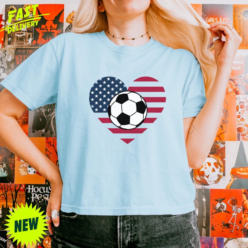 Puede incluir: Una camiseta de manga corta de color azul claro con un gr&aacute;fico en forma de coraz&oacute;n con los colores de la bandera estadounidense y un bal&oacute;n de f&uacute;tbol blanco y negro centrado en el medio. La prenda tiene cuello redondo y corte est&aacute;ndar.