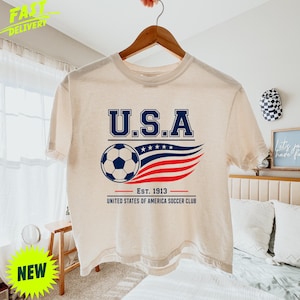 May include: A cream-colored short-sleeve t-shirt featuring a graphic with the text U.S.A. above a soccer ball and stylized red, white, and blue wavy lines. Below the graphic, the text reads EST. 1913 UNITED STATES OF AMERICA SOCCER CLUB.