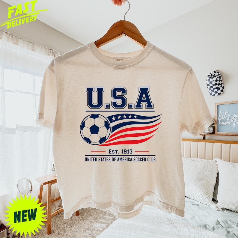 May include: A cream-colored short-sleeve t-shirt featuring a graphic with the text U.S.A. above a soccer ball and stylized red, white, and blue wavy lines. Below the graphic, the text reads EST. 1913 UNITED STATES OF AMERICA SOCCER CLUB.